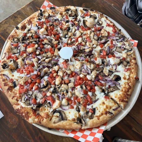 A large, round pizza topped with chicken, mushrooms, red onions, and diced tomatoes on a wooden table.