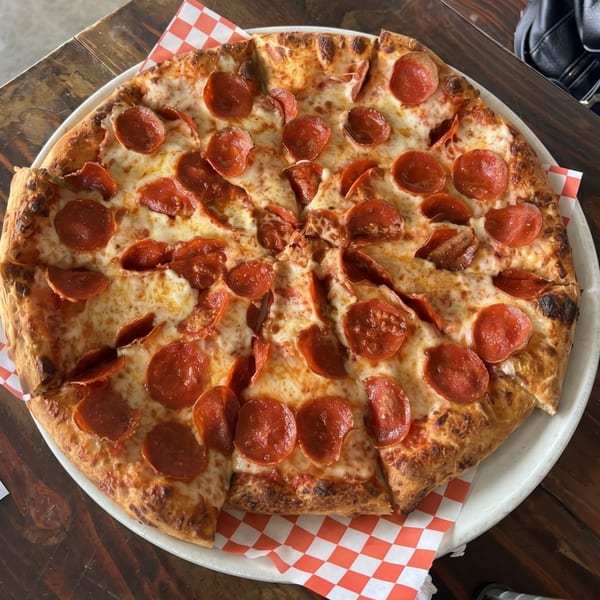 A whole pepperoni pizza, cut into slices, on a white plate with a red and white checkered napkin.