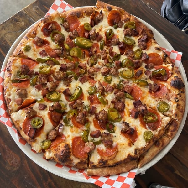 A whole pizza with pepperoni, sausage, and jalapeños on a white and red checkered paper.